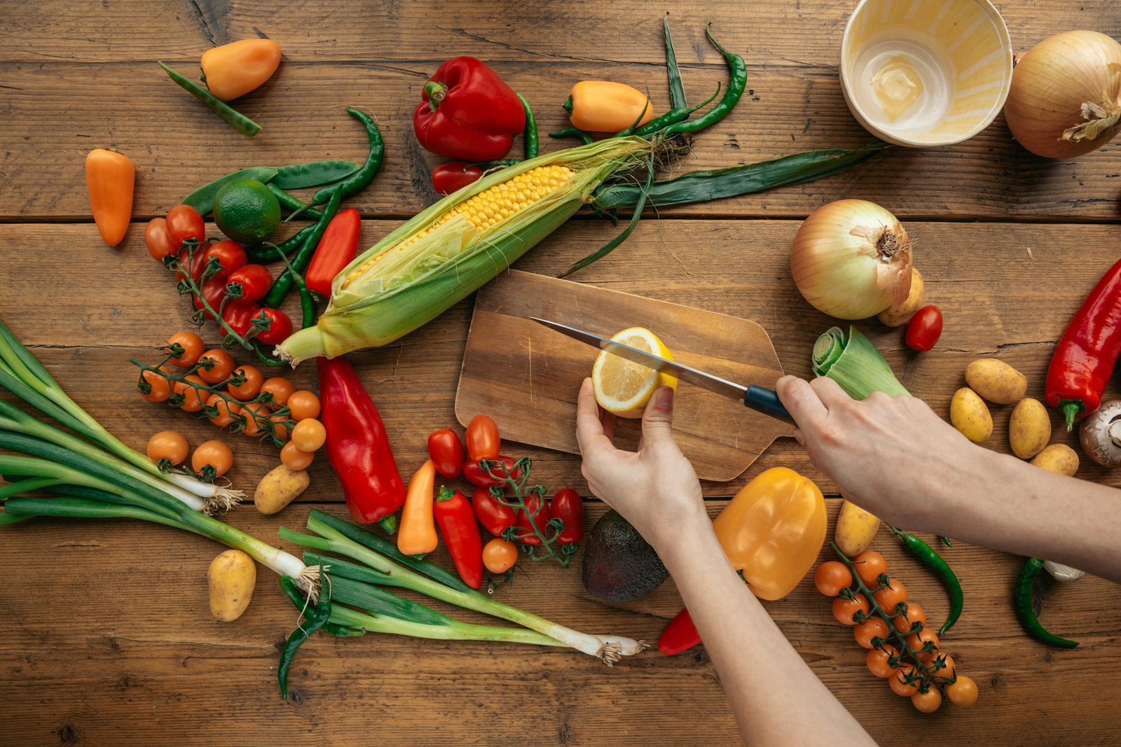 Top view of fresh vegetables and hands slicing a lemon on a wooden board.